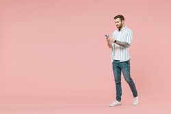 Full length side view young smiling unshaven man 20s in blue striped shirt hold mobile cell phone chat online browsing internet walk isolated on pastel pink background studio. Tattoo translate fun.