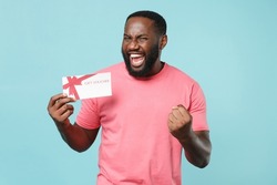 Joyful young african american man guy in casual pink t-shirt isolated on blue wall background studio portrait. People lifestyle concept. Mock up copy space. Hold gift certificate doing winner gesture