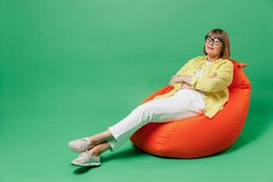 Full body elderly smiling minded happy woman 50s in glasses yellow shirt sit in bag chair resting relax during leisure time isolated on plain green background studio portrait People lifestyle concept