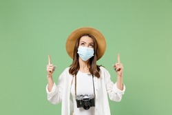 Traveler tourist woman in casual clothes hat face mask coronavirus covid19 lockdown point finger up on workspace isolated on green background Passenger travel abroad weekend Airflight journey concept