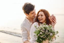 Young fun surprised couple family man woman in white clothes rest relax together boyfriend meet girlfriend close eyes gift give bouquet flowers at sunrise over sea beach outdoor seaside in summer day