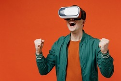 Smiling cheerful happy excited young brunet man 20s wear red t-shirt green jacket watching in vr headset pc gadget doing winner gesture celebrate isolated on plain orange background studio portrait.