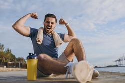 Full size young sporty strong toned fit sportsman man in sports clothes towel training near water bottle show biceps muscles at sunrise sun over sea beach outdoor on pier seaside in summer day morning