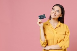 Young latin happy cute dreamful pensive wistful cute attractive satisfied rich successful woman 20s in yellow shirt hold credit bank card look aside isolated on pastel pink background studio portrait.