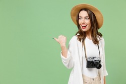 Traveler tourist woman in casual clothes hat camera point thumb finger back aside on workspace area isolated on green background Passenger travel abroad on weekends getaway. Air flight journey concept