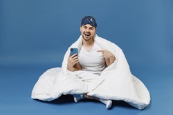 Full length happy young fun man in pajamas jam sleep mask rest relax at home sit wrap under cover blanket duvet point finger on mobile cell phone isolated on dark blue background Night bedtime concept