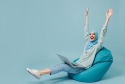 Full body young arabian asian muslim woman in abaya hijab sit in bag chair use work laptop pc computer stretch hands isolated on plain blue background People uae middle eastern islam religious concept