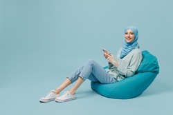 Full body happy young arabian asian muslim woman in abaya hijab sit in bag chair hold use mobile cell phone isolated on plain blue background studio People uae middle eastern islam religious concept