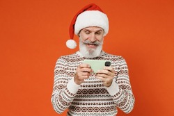 Old happy bearded Santa Claus man 50s wears Christmas hat sweater posing using play racing app on mobile cell phone hold gadget smartphone for pc video games isolated on plain orange background studio