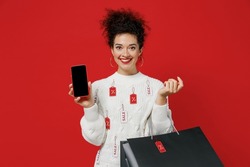 Young fun happy woman in white sweater with tags sale hold package bags with purchases after shopping using mobile cell phone with blank screen workspace area isolated on plain red background studio.