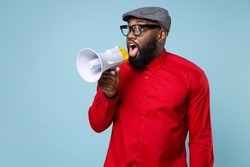Shocked amazed young bearded african american man 20s wearing casual red shirt eyeglasses cap standing screaming in megaphone looking aside isolated on pastel blue color background studio portrait