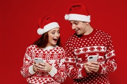 Young shocked couple friends two man woman in sweater hat hold in hand use mobile cell phone isolated on plain red background studio. Happy New Year 2022 celebration merry ho x-mas holiday concept.