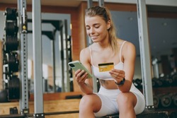 Young strong sportswoman woman wear white sportswear warm up train sit near trainers using mobile cell phone credit bank card for online shop pay gym membership in gym indoors Workout sport concept.