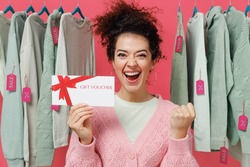 Young overjoyed woman in sweater stand near clothes rack with tag sale in store showroom hold gift certificate coupon voucher card for store do winner gesture isolated on plain pink background studio