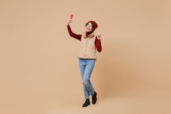 Full body young woman wears red turtleneck vest beret doing selfie shot on mobile cell phone post photo on social network show victory sign isolated on plain pastel beige background studio portrait.