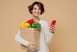 Young happy fun vegetarian woman 20s in casual clothes hold paper bag with vegetables hold in hand use mobile cell phone read browsing menu isolated on plain pastel beige background Shopping concept