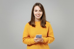 Young smiling cheerful woman 20s wearing casual knitted yellow sweater hold in hand using mobile cell phone chatting browsing typing sms messaging isolated on grey color background studio portrait
