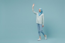 Full body fun young arabian asian muslim woman in abaya hijab hold hands crossed folded walk go waving hand isolated on plain blue background studio People uae middle eastern islam religious concept.