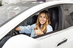 Happy woman customer buyer client in blue shirt hold put hands on steering wheel show thumb up choose auto want buy new automobile in car showroom vehicle salon dealership store motor show indoor.