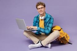 Full size young boy teen student in casual clothes backpack headphones glasses sit hold work on laptop computer isolated on violet background studio Education in high school university college concept