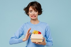 Young woman wear sweater hold a box with fastfood french fries potato burger show stop palm gesture reject say no isolated on plain pastel light blue background. People lifestyle junk food concept.