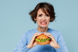 Young sad irritated stressed woman 20s wearing casual sweater look camera biting eating burger isolated on plain pastel light blue color background studio portrait. People lifestyle junk food concept