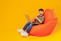 Full body little small fun boy 6-7 years old in green t-shirt sit in bag chair hold use work point on laptop pc computer isolated on plain yellow background Mother's Day love family lifestyle concept