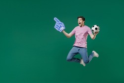 Full size body length young fun man fan wear pink t-shirt cheer up support football sport team hold in hand soccer ball watch tv live stream scream jump isolated on dark green color background studio