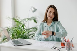 Young smiling happy successful employee business woman 20s in blue shirt look at smartwatch time sit work at workplace white desk with laptop pc computer at office indoors. Achievement career concept