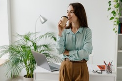 Young minded successful employee business woman in blue shirt work hold takeaway delivery craft paper cup coffee to go stand at desk with laptop pc computer at office indoor Achievement career concept