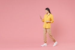 Full length side view of young smiling brunette positive nice attractive latin woman 20s wearing yellow shirt hold mobile cell phone walking isolated on pastel pink color background studio portrait