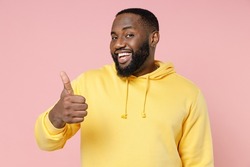 Smiling cheerful funny young african american man 20s wearing casual basic yellow streetwear hoodie standing showing thumb up looking camera isolated on pastel pink color background studio portrait