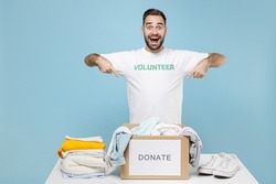 Shocked young man in volunteer t-shirt stand near table pointing index fingers on donation box clothes for needy isolated on blue background. Voluntary free work assistance help charity grace concept