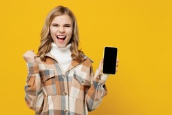 Little smiling surprised happy blonde kid girl 13-14 years in checkered shirt hold in hand use mobile cell phone with blank screen workspace area do winner gesture isolated on plain yellow background