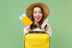 Close up excited tourist woman in casual clothes hat hold passport tickets yellow suitcase spread hands isolated on green background Passenger travel abroad weekend getaway Air flight journey concept.