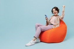 Full body young smiling cheerful cool happy woman 20s wear brown shirt sit in bag chair hold use mobile cell phone do winner gesture isolated on pastel plain light blue background studio portrait.