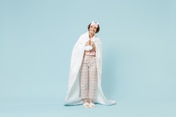 Full length young fun woman 20s in pajamas jam sleep eye mask rest relax at home wrap covered under blanket duvet look aside isolated on pastel blue background studio Good mood night bedtime concept
