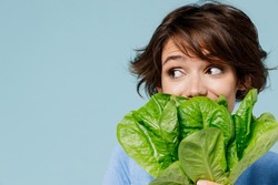 Close up young woman 20s in sweater hold cover mouth with green lettuce leaves look aside on workspace isolated on plain pastel light blue background studio portrait. People lifestyle food concept.