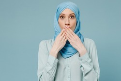 Young shocked surprised astonished arabian asian muslim woman in abaya hijab cover mouth with hand isolated on plain blue background studio portrait. People uae middle eastern islam religious concept