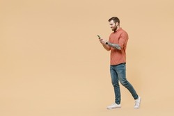 Full size body length side view happy tatooed young brunet man 20s short haircut wears apricot shirt go stride hold in hand use mobile cell phone isolated on pastel orange background studio portrait.