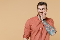 Worried sad tatooed young brunet man 20s short haircut with earrings wears apricot shirt look aside bite nails isolated on pastel orange background studio portrait. People emotions lifestyle concept.