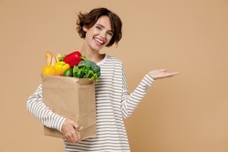 Young happy vegetarian woman 20s in casual clothes hold paper bag with vegetables point hand arm aside on workspace area copy space mock up isolated on plain pastel beige background Shopping concept