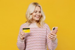 Elderly smiling blonde woman 50s in pink sweater using mobile cell phone hold credit bank card doing online shopping order delivery booking tour isolated on plain yellow background studio portrait.
