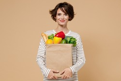 Young smiling caucasian cheerful happy fun vegetarian woman 20s in casual clothes hold paper bag with vegetables after shopping look camera isolated on plain pastel beige background studio portrait