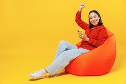 Full size body length young woman of Asian ethnicity 20s in casual clothes sit in bag chair hold in hand use mobile cell phone doing winner gesture isolated on plain yellow background studio portrait