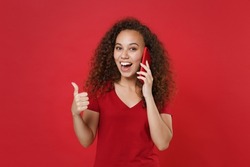 Excited young african american girl in casual t-shirt posing isolated on red wall background studio portrait. People lifestyle concept. Mock up copy space. Talking on mobile phone, showing thumb up