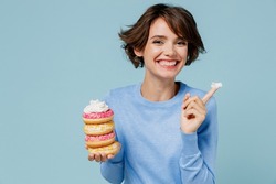 Young happy woman 20s wearing casual sweater look camera hold donuts dessert show finger with cream isolated on plain pastel light blue background studio. People lifestyle junk unhealthy food concept
