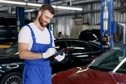 Young minded professional car mechanic man in denim blue overalls white t-shirt gloves hold clipboard with papers document work in modern vehicle repair shop workshop indoors. Tattoo translate fun.