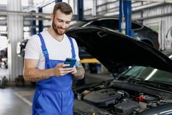 Young professional technician car mechanic man 20s in blue overalls white t-shirt use hold mobile cell phone browsing fix problem with raised hood bonnet work in vehicle repair shop workshop indoor.