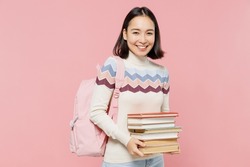 Smiling happy fun teen student girl of Asian ethnicity wearing sweater backpack hold pile of books look camera isolated on pastel plain light pink background Education in university college concept